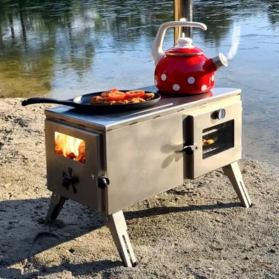 A stainless steel outdoor stove with a red and white polka dot kettle and a pan of food on top, set up by the water's edge.