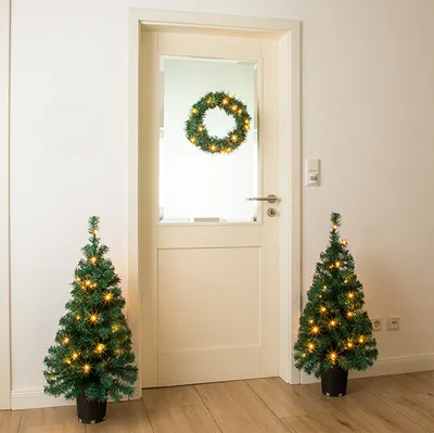 Two small, lit Christmas trees flank a door with a lit wreath on it.
