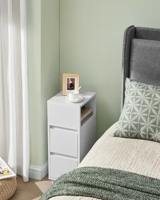 A white bedside table with drawers next to a bed with a grey headboard and a patterned pillow.