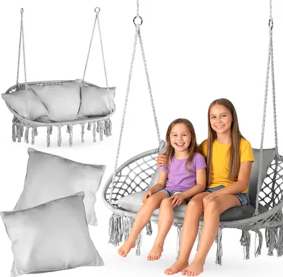 Two young girls sitting in a hanging chair swing, with another hanging chair and pillows in the background.