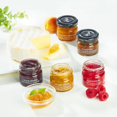 A cheese board setup with a wheel of brie cheese, a slice of cheese, and several small jars of preserves, along with fresh raspberries and a sprig of mint.