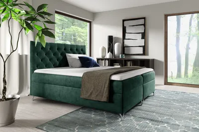 A bedroom scene with a green boxspring bed, large windows overlooking a forest, and decorative elements.