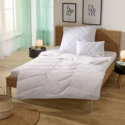 A bedroom scene with a neatly made bed featuring a white duvet and pillows, set against a woven headboard. The room has light wooden flooring and sheer green curtains.