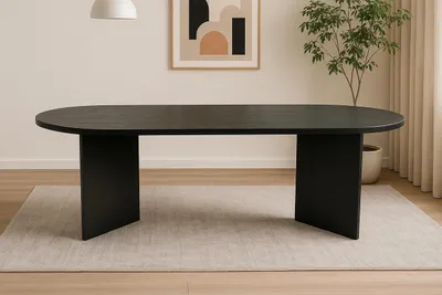 A black oval dining table with V-shaped legs is placed on a light gray rug in a well-lit room. A framed abstract artwork hangs on the wall behind the table, and a potted plant is visible to the right.