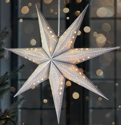 A silver 3D paper star with small lights is hanging in front of a window with bokeh lights.