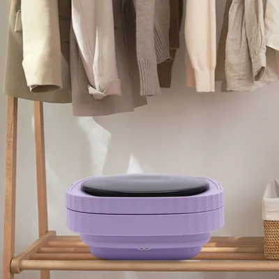 A purple, foldable mini washing machine with a black lid sits on a wooden shelf. Clothes hang on a rack above it.