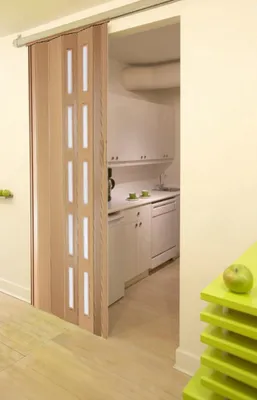 A folding door is partially open, revealing a small kitchen area with white cabinets and appliances. A bright green shelf with an apple is in the foreground.