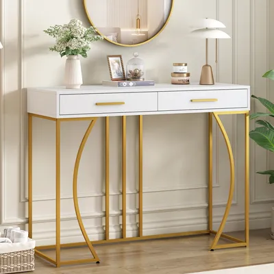 A white and gold console table with two drawers, decorated with a vase of flowers, a framed picture, and a lamp.