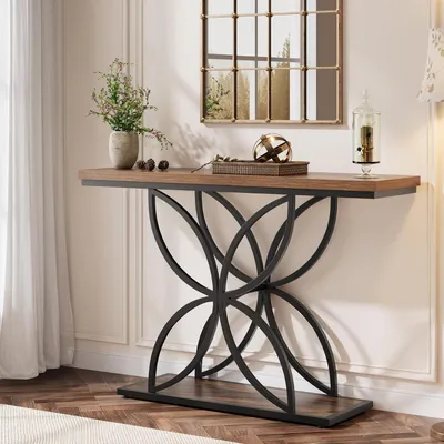 A console table with decorative items is placed against a wall with molding and a mirror above it. A window with sheer curtains is to the left.