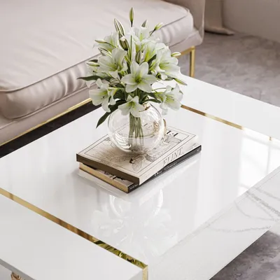 A close-up of a white high-gloss coffee table with gold accents, featuring a vase of white lilies and two books.