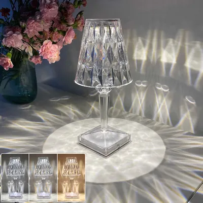 A clear acrylic table lamp is shown on a surface, casting intricate light patterns on the wall and surface. A vase of pink flowers is visible in the background. Three smaller inset images show the lamp in different light color settings.