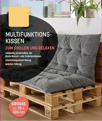 A large cushion is placed on wooden pallets, suggesting an outdoor or casual seating arrangement. The cushion is shown in a reclined position, as if for relaxation.