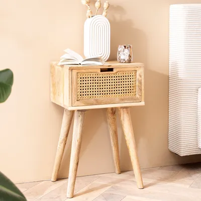 A wooden nightstand with a rattan drawer front, decorated with a vase and a book, placed next to a bed.
