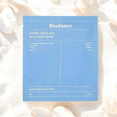 A blue package for a face mask is laid out on a sandy surface with seashells.