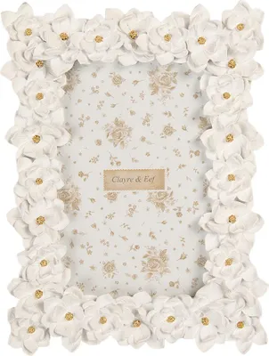 A white picture frame decorated with fabric flowers and gold accents, set against a floral patterned background.