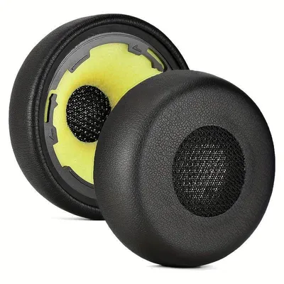 Two black ear pads for headphones are shown against a white background. One ear pad is shown from the side, revealing its internal yellow foam and black mesh speaker cover. The other ear pad is shown from the front, also with a black mesh speaker cover.