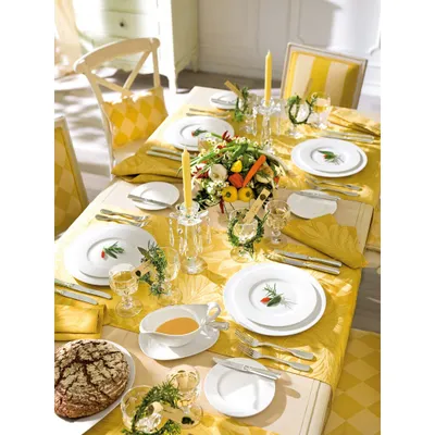 A beautifully set dining table with a yellow tablecloth, white plates, cutlery, candles, and a centerpiece of vegetables and greenery.