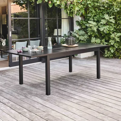 A dark aluminum extendable garden table is set on a wooden deck, with lush green foliage and a glass door in the background.