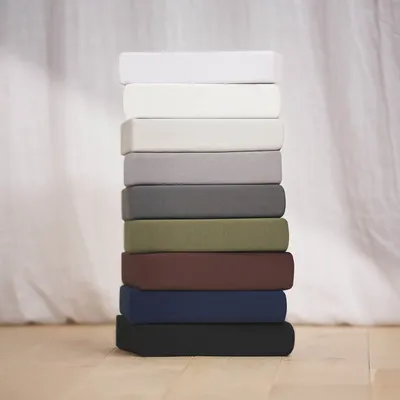 A stack of fitted sheets in various colors, arranged vertically against a white, textured background.