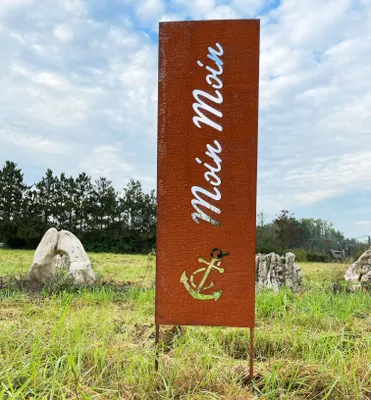 A tall, rectangular metal sign with "Moin Moin" and an anchor design, standing in a grassy field with trees and rocks in the background.