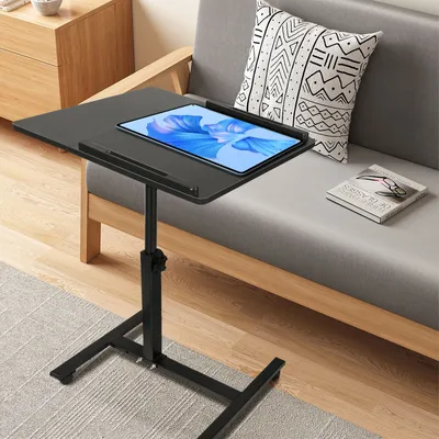 A black adjustable laptop table with a tablet on it, placed next to a gray sofa with a patterned pillow and a book.