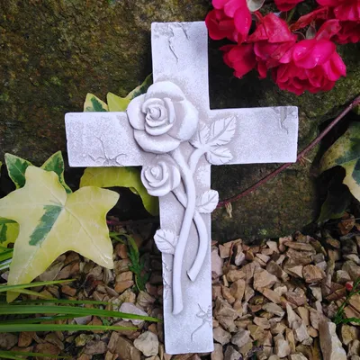 A close-up of a white cross with a 3D rose design, placed outdoors among rocks, ivy, and pink flowers.