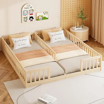 Two children's beds are placed side-by-side in a room with a light-colored rug and wooden floor. There are decorative pictures on the wall and a small side table with a plant and books.