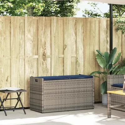 An outdoor storage bench with a cushion sits on a patio next to a wooden fence and a small folding table.