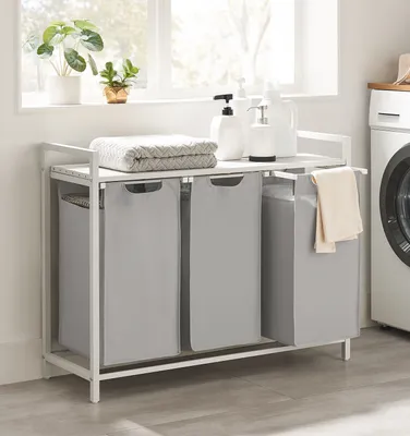 A laundry basket with three compartments and a shelf on top, placed in a laundry room setting.