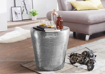A silver hammered metal side table is placed next to a sofa in a living room. A miniature motorcycle model is on the rug in front of the table.