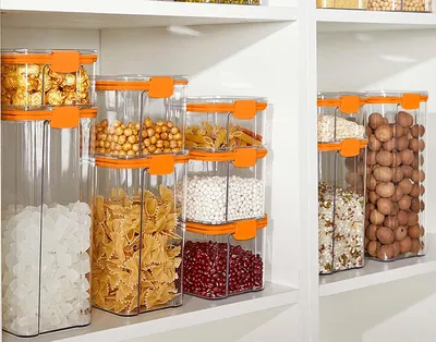 A collection of clear plastic food storage containers with orange lids, filled with various dry goods like pasta, beans, and potatoes, neatly arranged on white shelves.
