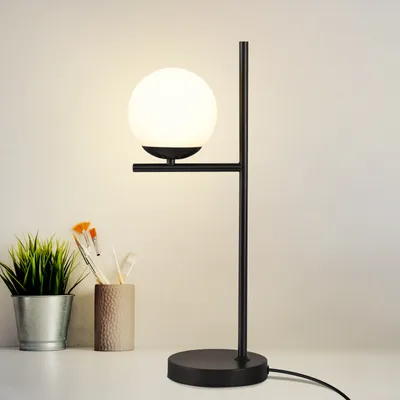 A modern black desk lamp with a white glass shade sits on a white surface next to a potted plant and a holder with brushes.