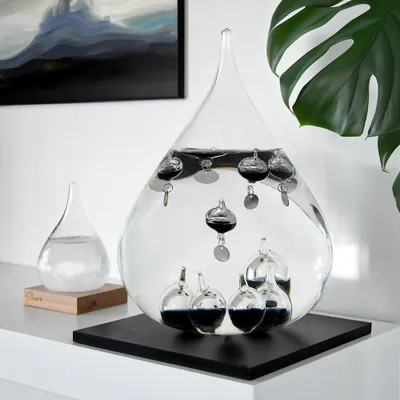A Galileo thermometer is displayed on a shelf next to a smaller glass container, with a plant and artwork in the background.