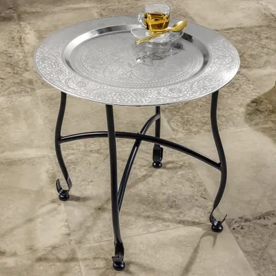 A round silver side table with a decorative embossed pattern on the tray top, featuring a cup of tea and a spoon. The table has black metal legs and is placed on a tiled floor.
