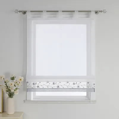 A white sheer roman blind with embroidered floral detail is shown hanging on a window.
