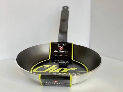A De Buyer Choc Access frying pan is displayed against a white background. The pan has a black handle and a silver-grey interior and exterior. A yellow and black label is wrapped around the pan, featuring the brand name and product details.