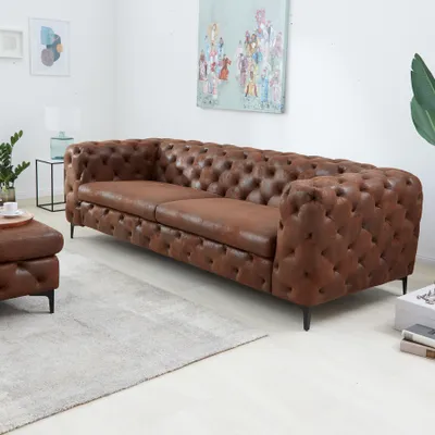 A brown Chesterfield sofa and ottoman are shown in a living room setting with a rug and artwork on the wall.