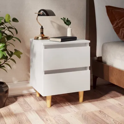 A white bedside table with two drawers, a lamp, a book, and a small plant on top, placed next to a bed.