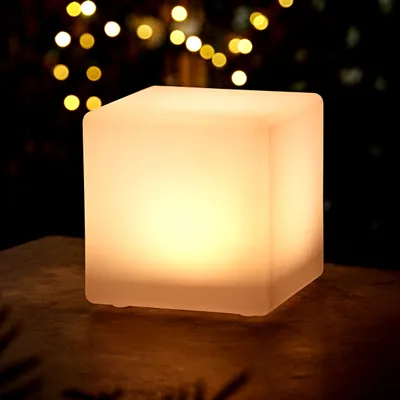 A glowing cube-shaped lamp sits on a textured surface, with blurred lights in the background.