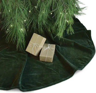 A close-up shot of a dark green velvet Christmas tree skirt with two brown wrapped gifts tied with white string, placed under pine branches with fairy lights.