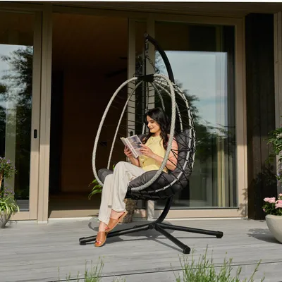 A woman sits in a hanging chair on a deck, reading a book.