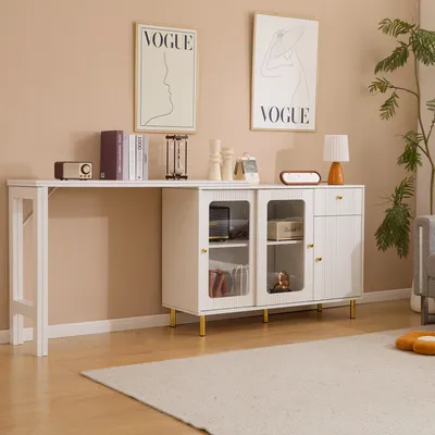 A white sideboard with glass doors and gold legs is placed against a light beige wall, with a long white table attached to its left. Two framed Vogue posters hang on the wall above the furniture. A large rug covers the wooden floor in front of the sideboar