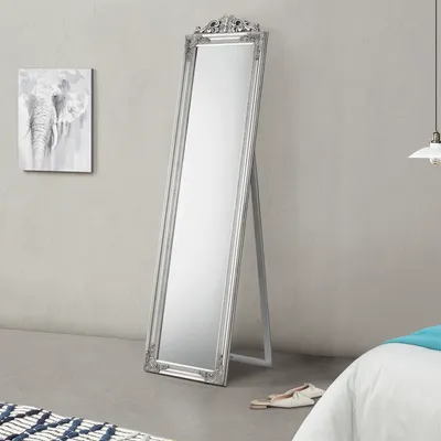 A full-length silver mirror with an ornate frame stands on a grey floor against a light grey wall. A painting of elephants hangs on the wall to the left, and a white bed with a blue throw is visible on the right.