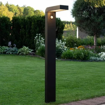 A modern black solar-powered garden light with a motion sensor is illuminated in a lush green garden with various plants and trees in the background.