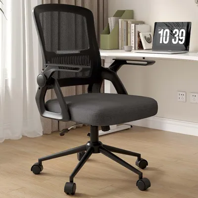 A black mesh office chair is shown in a home office setting with a desk and laptop in the background.