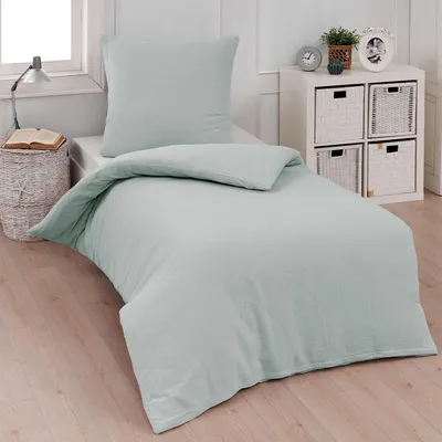 A neatly made bed with a single pillow and duvet cover in a light silver-grey color, set in a bedroom with a bedside table and storage unit.