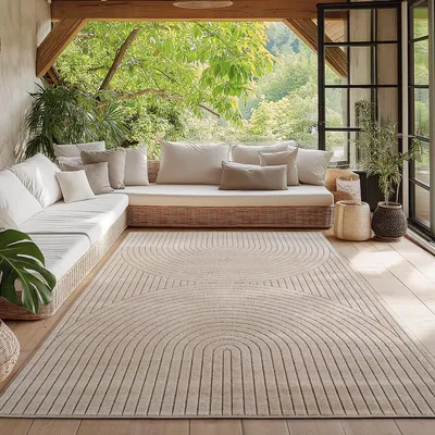 An outdoor rug is placed on a wooden floor on a covered patio, with a large sectional sofa and lush greenery visible in the background.