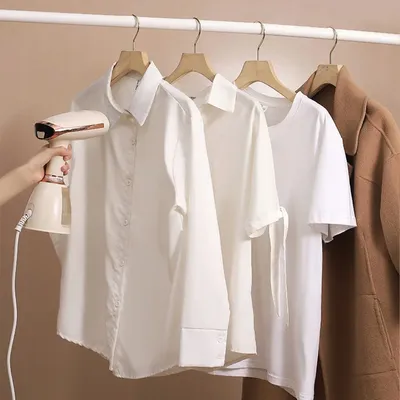 A hand holds a beige and pink handheld steamer near white shirts hanging on a rack, with a brown coat on the far right.