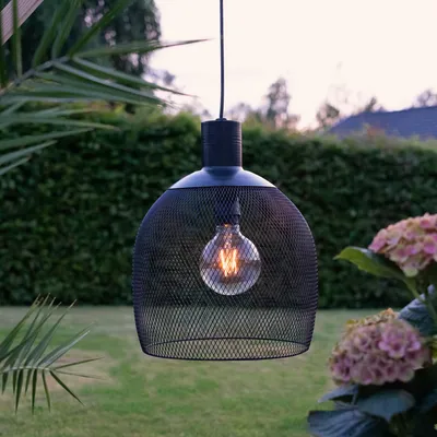A black pendant lamp with a mesh shade hangs outdoors in a garden setting, with lush greenery and flowers in the background.