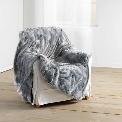 A plush gray faux fur blanket draped over a white armchair in a room with light wood floors and a window with sheer curtains.